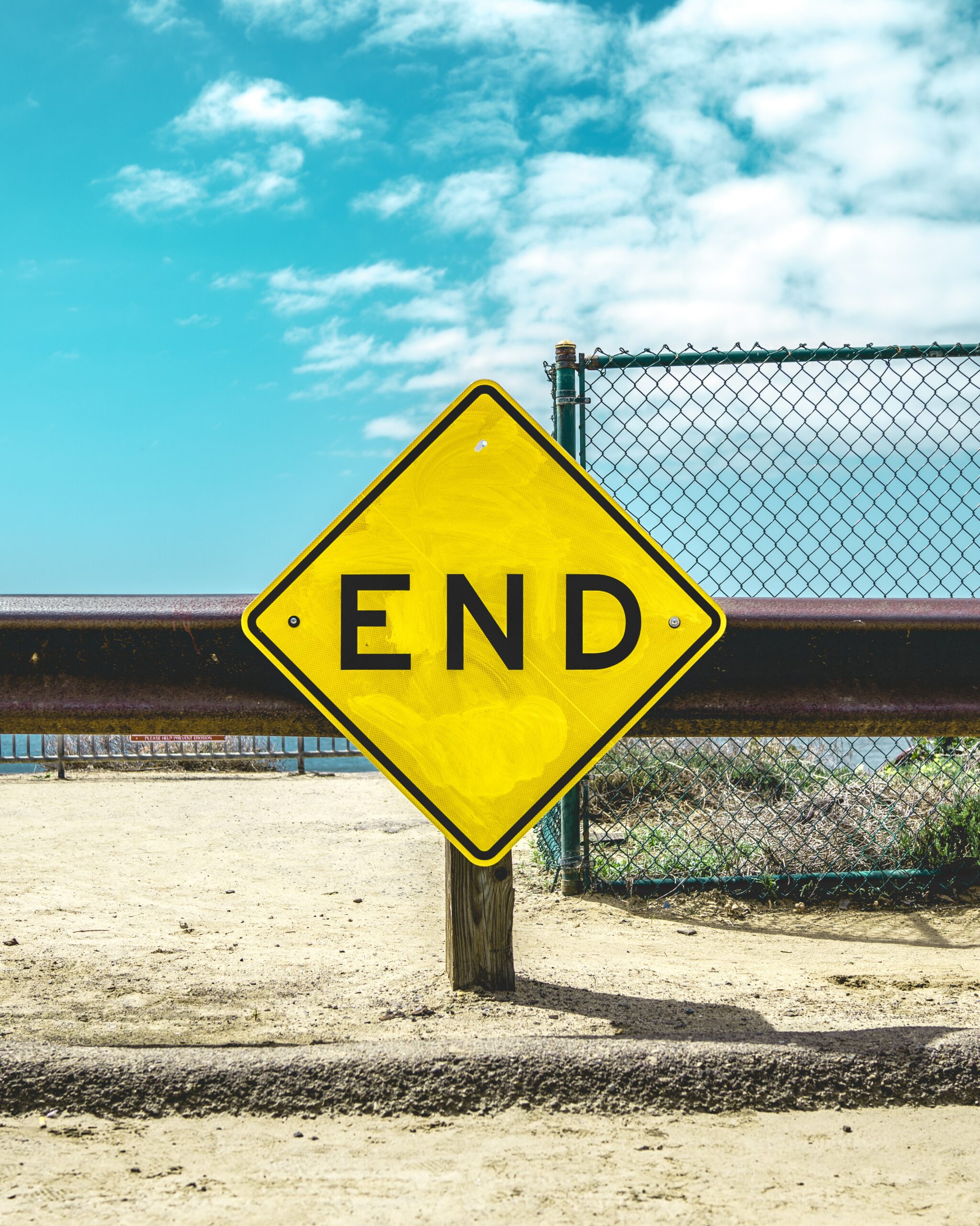 A yellow diamond sign says END against a blue and cloudy sky. It is planted in what appears to be dry dirt or sand and there is a chain-link fence behind it.